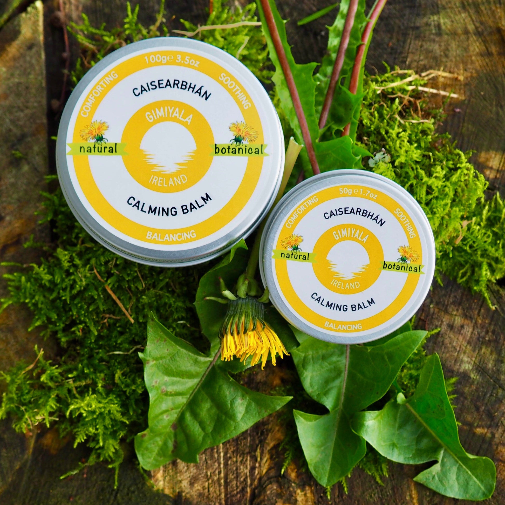 Two tins of Calming Balm on a wooden surface with green leaves and plants.